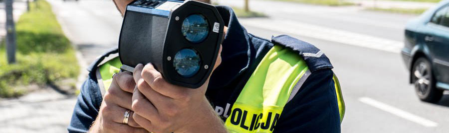 zdjęcie przedstawia policjanta drogówki z radarowym miernikiem prędkości w trakcie dokonywania pomiaru