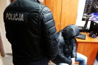 zdjęcie - dwie osoby, jedna stoi w kamizelce z napisem policja, druga siedzi w czarnej kurtce i kapturze na głowie