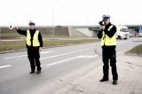 Dwóch policjantów drogówki, jeden z tarczą zatrzymuje pojazd, drugi dokonuje pomiaru prędkości laserowym miernikiem prędkości.