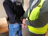 Zdjęcie - policjant w kamizelce odblaskowej z napisem POLICJA i zatrzymany w kajdankach