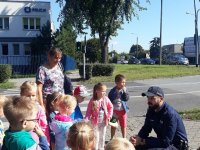 Policjant tłumaczy dzieciom jak zachować się na przejściu dla pieszych - na zdjęciu dzieci z przedszkola i policjant w tle ulica i budynek komisariatu