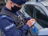 zdjęcie poglądowe - policjant stojący przy radiowozie z notatnikiem w ręku, twarz ma zasłoniętą maseczką