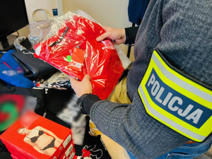 zdjęcie - policjant trzyma w ręku podrobione polo, a na biurku leżą inne towary z podrobionymi znakami towarowymi